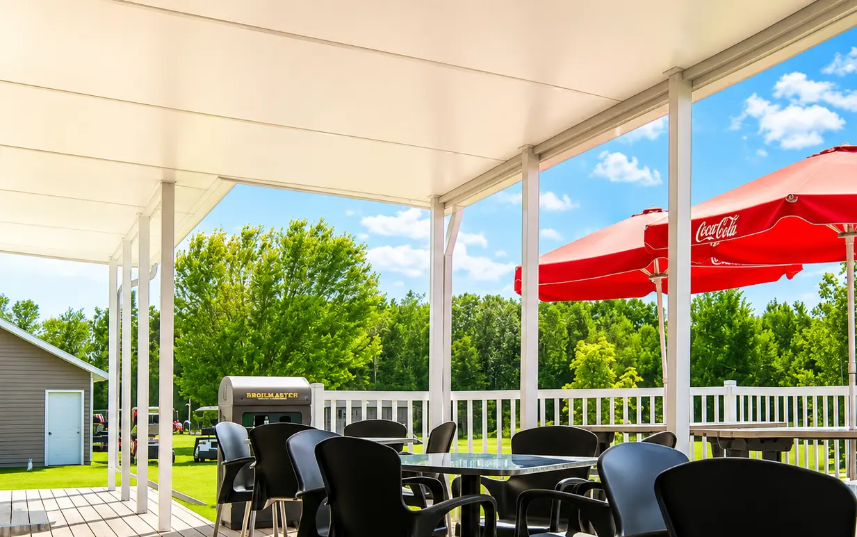 View under a white solid insulated patio cover, featuring a spacious outdoor dining area with black chairs and red patio umbrellas, overlooking a grassy yard with trees.