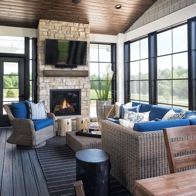A beautifully designed sunroom with a stone fireplace, blue lounge furniture, and floor-to-ceiling black-framed windows overlooking a green yard.