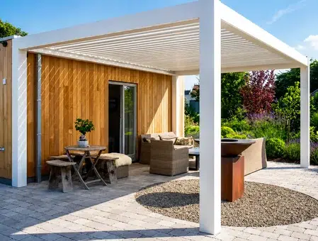 A modern white louvered roof pergola covering a stone patio with outdoor sofas, a small wooden table, and a fire pit next to a wood-paneled wall.